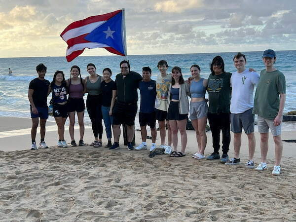 Students stand on the beach against blue water holding up the Puerto Rico flag