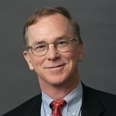 A headshot of a man wearing glasses, a dark suit jacket, light blue collared shirt, and patterned red tie. He smiles slightly against a gray background.