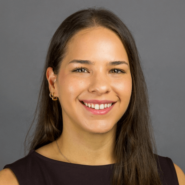 Headshot of a young woman with long brown hair, wearing a dark sleeveless top, smiling against a gray background.