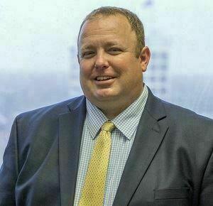 Man in a navy suit and gold tie smiles against a blurred background.