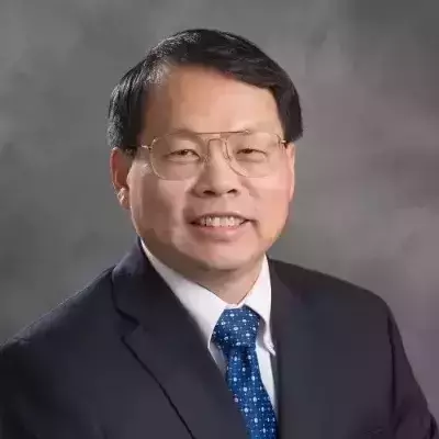 A smiling man with glasses in a dark suit, white shirt, and blue patterned tie against a grey background.