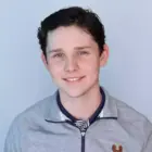 Young man with dark hair and blue eyes smiles, wearing a grey zip-up jacket over a navy and white striped polo.