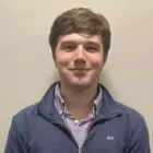 Young man with light brown hair wearing a blue quarter-zip sweater and purple checkered shirt, smiling slightly.