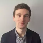 Young man with curly brown hair, blue eyes, smiling. Wears a navy blazer over a black and white gingham shirt.