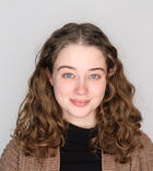 Smiling young woman with light brown curly hair and blue eyes, wearing a black shirt under a textured brown cardigan.