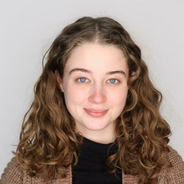 Smiling young woman with light brown curly hair and blue eyes, wearing a black shirt under a textured brown cardigan.