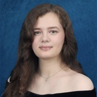Young woman with long brown hair, light eyes, and a subtle smile wears a dark off-the-shoulder top and silver necklace.