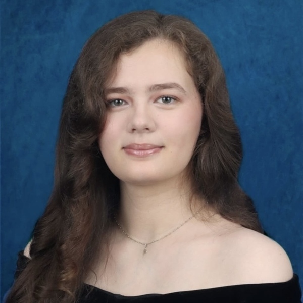 Young woman with long brown hair, light eyes, and a subtle smile wears a dark off-the-shoulder top and silver necklace.