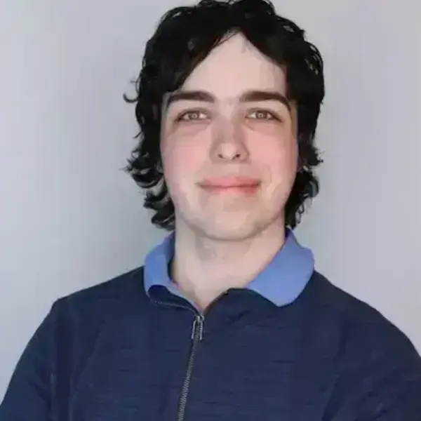 Young man with dark, curly hair and light eyes smiling, wearing a dark blue zip-up jacket over a light blue collar.