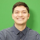 Young man with short brown hair smiles broadly, wearing a navy polka-dot shirt against a green background.