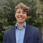Smiling man with curly brown hair, light blue shirt, navy blazer, and green foliage background.