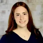 A young woman with long reddish-brown hair, smiling, wearing a dark blue top, and a small silver heart pendant necklace.