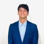 Smiling young man with dark curly hair, wearing a navy blue suit jacket and light blue collared shirt.